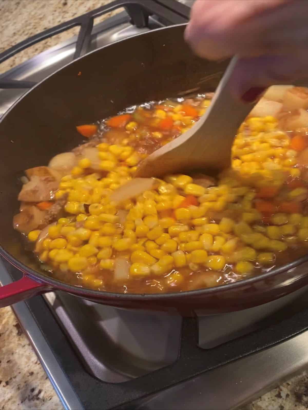 Stirring corn into a broth full of potatoes, carrots, celery, onion,and sausage