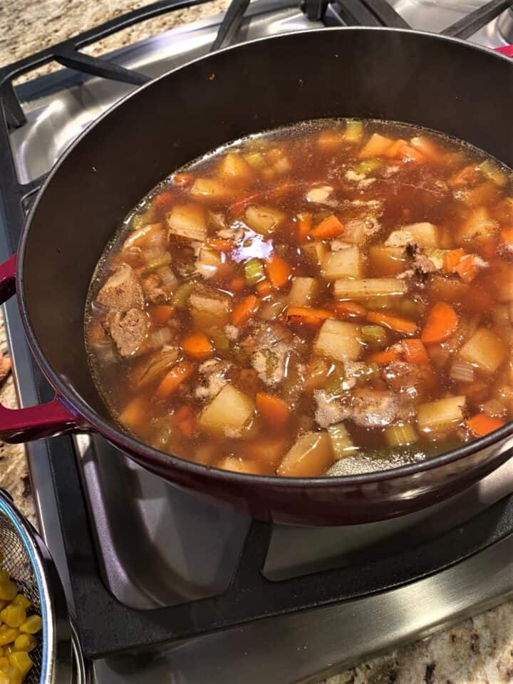 Cooked sausage crumbles, potato cubes, celery, carrot, and onion bits float in a brownish broth in Dutch oven on a gas burner, Colander of corn on the side