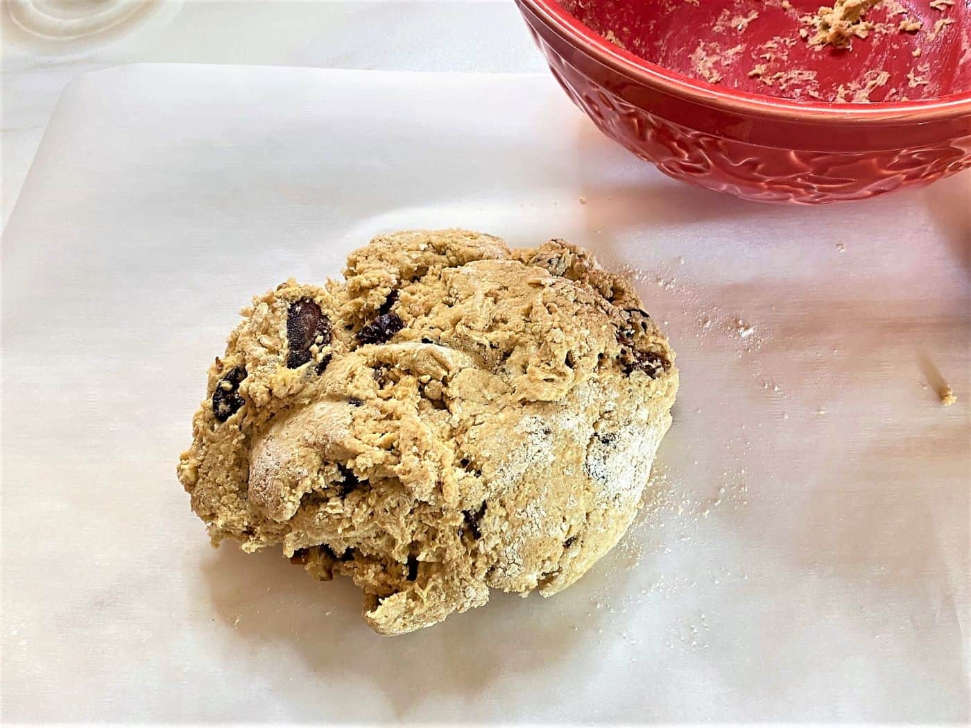 Roundish blob of dough studded with dates on white floured parchment and red mixing bowl in the right corner