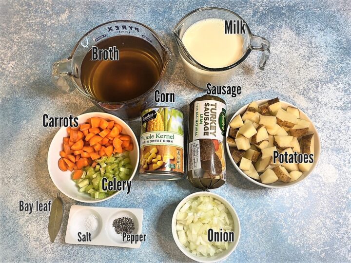 Onion, salt crystals, black pepper, bay leaf, carrot, celery, cubed potatoes with skins on, tube of turkey sausage, can of corn, broth, and milk on a bluish backgroud: Labelled ingredients for Corn and Sausage Chowder