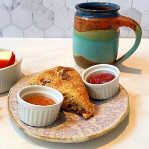 Pottery mug with vibrant southwest colors, wedge scone on small plate flanked by small white ceramic round dishes of jam on marble countertop