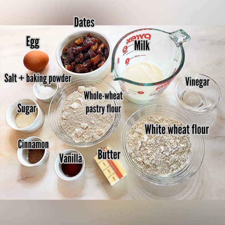 ingredients for date scones shown measured in clear glass and white porcelain containers plus a measuring cup with milk and half a stick of butter on a white marble background