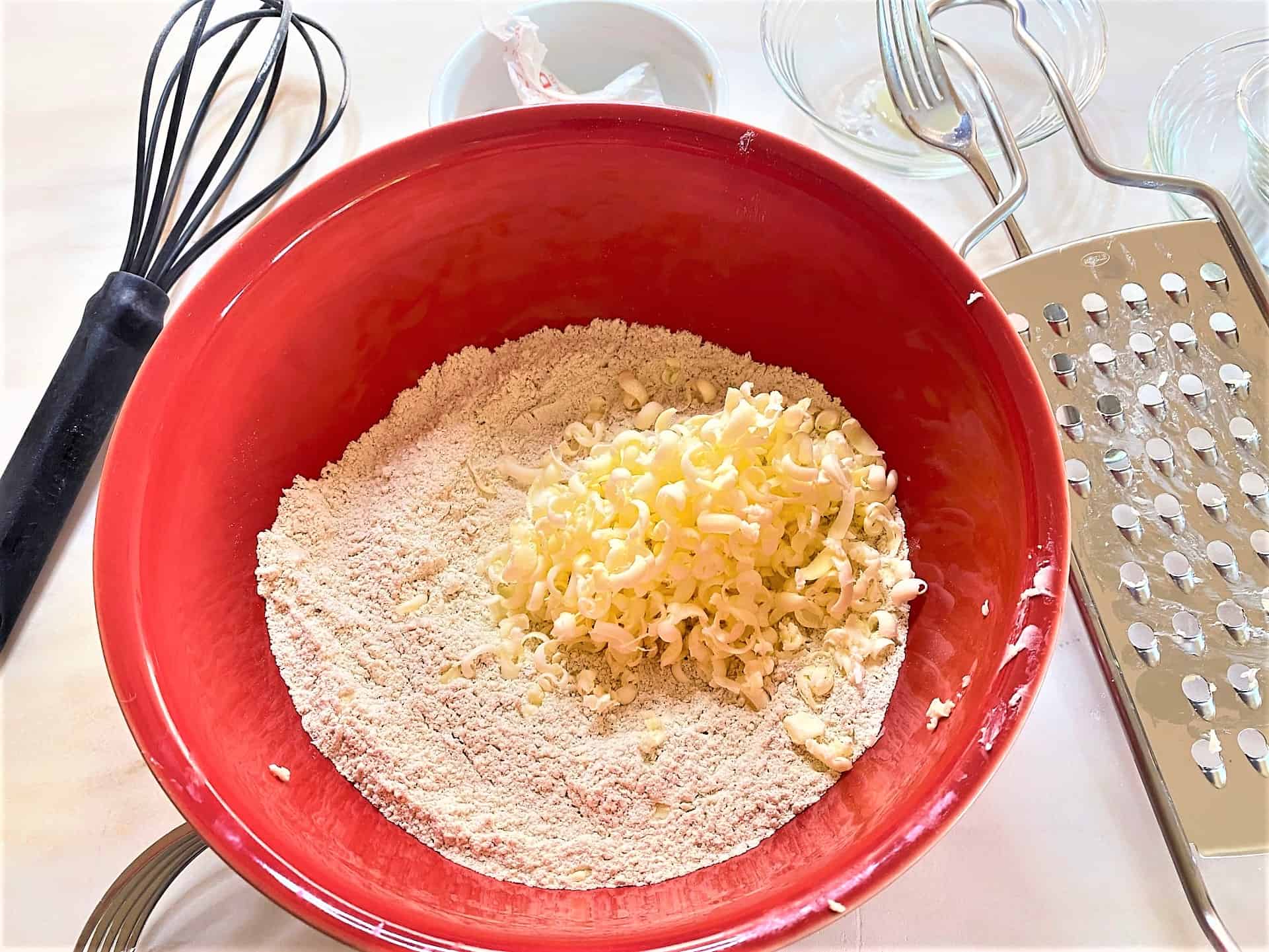 Shreds of grated yellow butter on top of flours in red bowl with whisk off to the side and grater