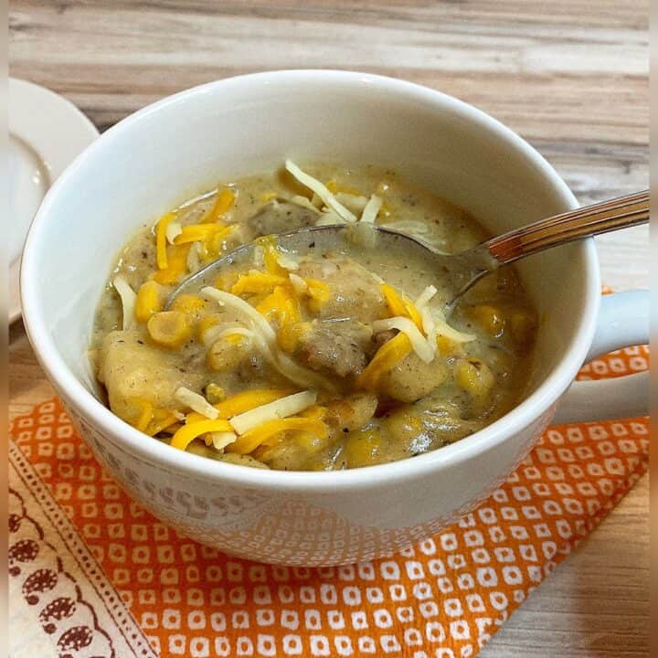 White bowl full of amber colored soup containing corn, bulk sausage bits, and potatoes with skins, with shreds of orange and white cheese flotaing on top. Spoon sticking bowl on colorful orange-white patterned napkin