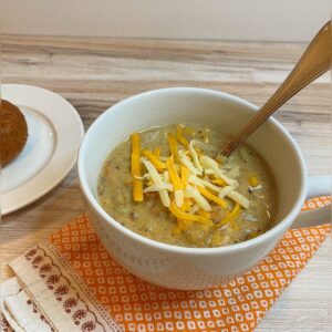 White bowl with thick amber soup topped with orange and white shreds of cheese in a white bowl with a spoon sticking out on a colorful orange white patterned cloth napkin with a small white plate in upper left corner with a brown edge of a roll. On a wood-grain counter top