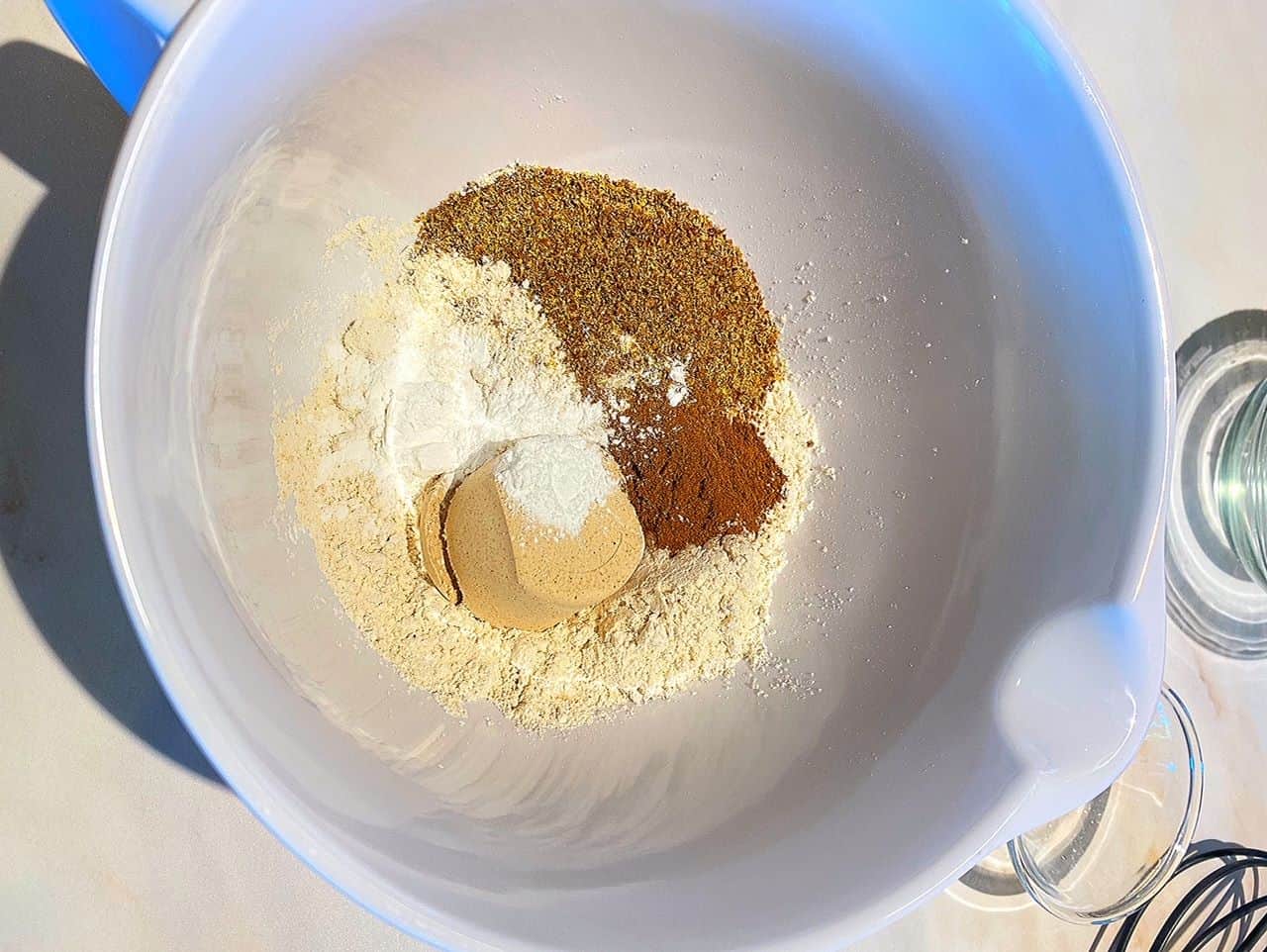 Cream-colored flour and protein powder with brown flaxseed, maroon spices, and white salt and baking powder in a white melamine spouted bowl