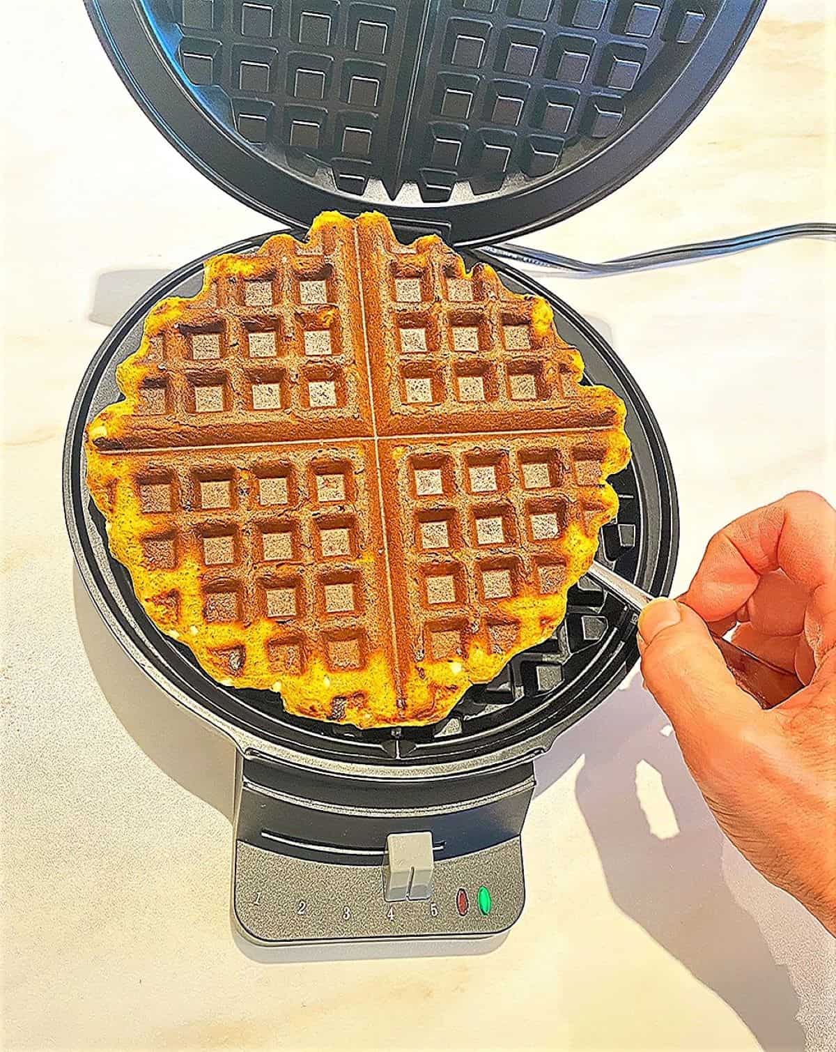 Golden yellow-brown cooked waffle with four sections and grid marks being taken out of a round wafflemaker with black grids