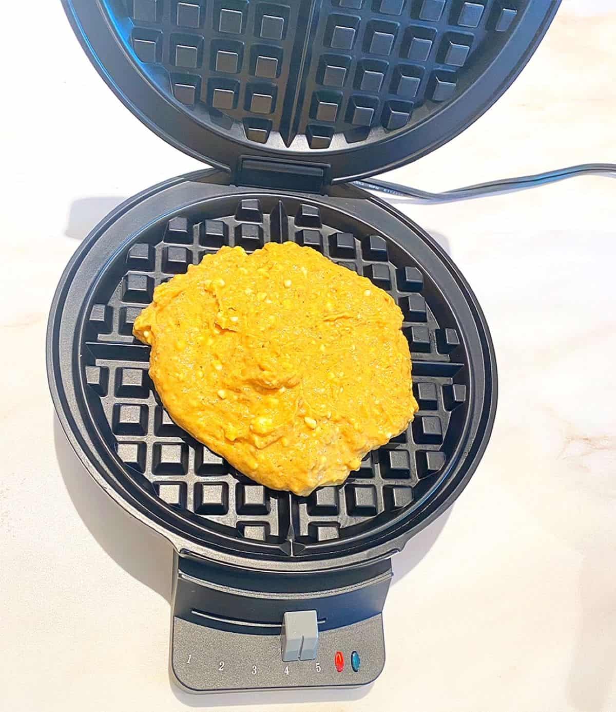 Ragged circle of thick, deep yellow, lumpy batter on a round waffle grid on a white marble countertop