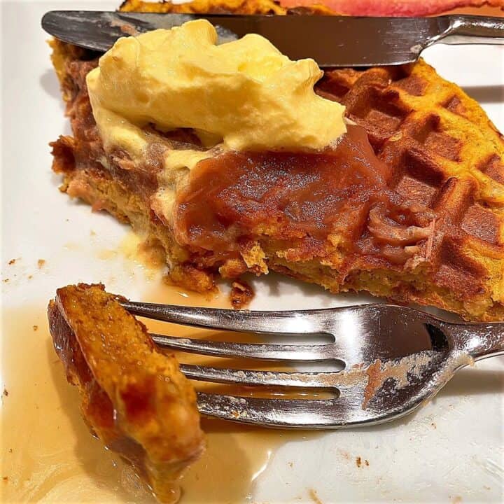 Fork with a bit of pumpkin waffle covered in syrup next to half-eaten waffle with whipped cream and apple butter