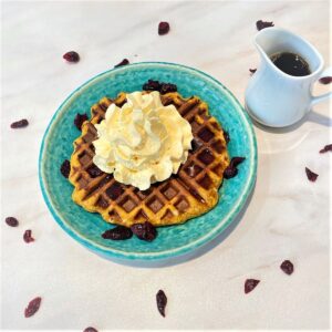 Round, ragged edge waffle topped with whipped cream and syrup on a round blue-green plate with a small white pitcher full of brown syrup next to it on a whitish marble countertop sprinkled with craisins