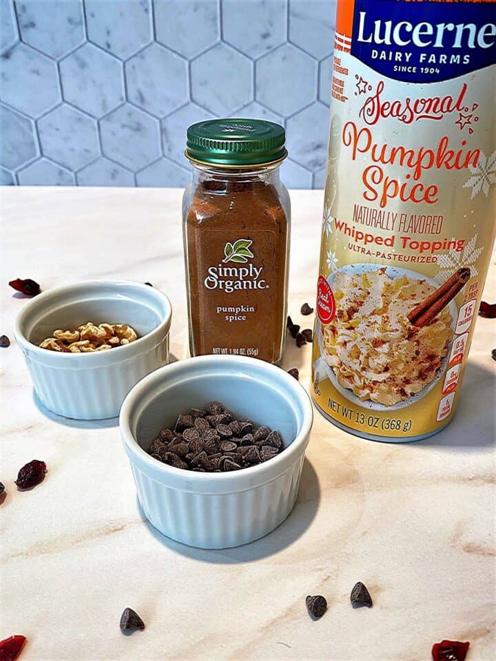 Can of pumpkin spice whipped cream, jar of pumpkin spice, small white round dishes with walnuts and mini chocolate chips on light marble countertop with grayish hex tile backsplash