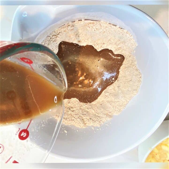 Pouring brownish liquid ingredients into dry ingredients in a white bowl to make batter for apple pear bread.