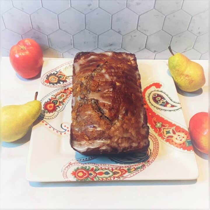 Rectangular loaf of deep golden apple pear bread on a paisley plate with fresh whole pears and apples next to the plate.