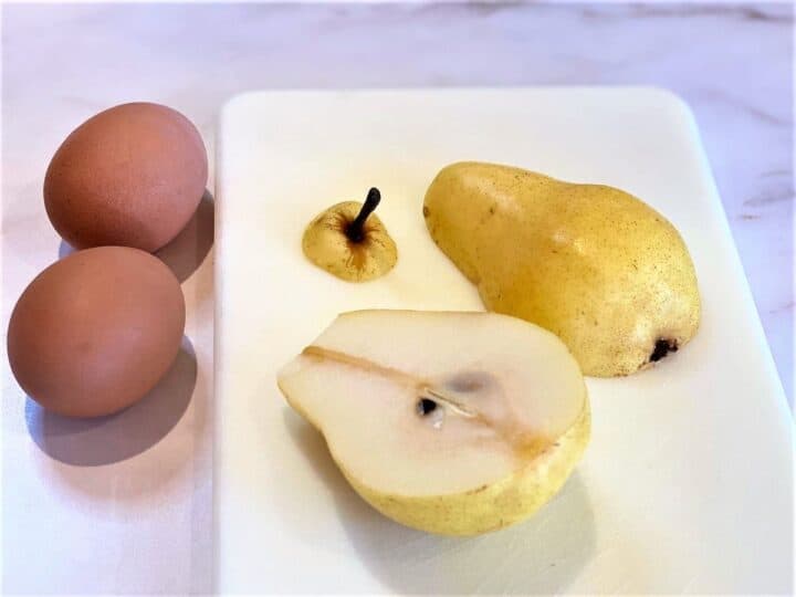 Bartlett pear with yellow skin but in half on white cutting board with a couple of brown eggs.