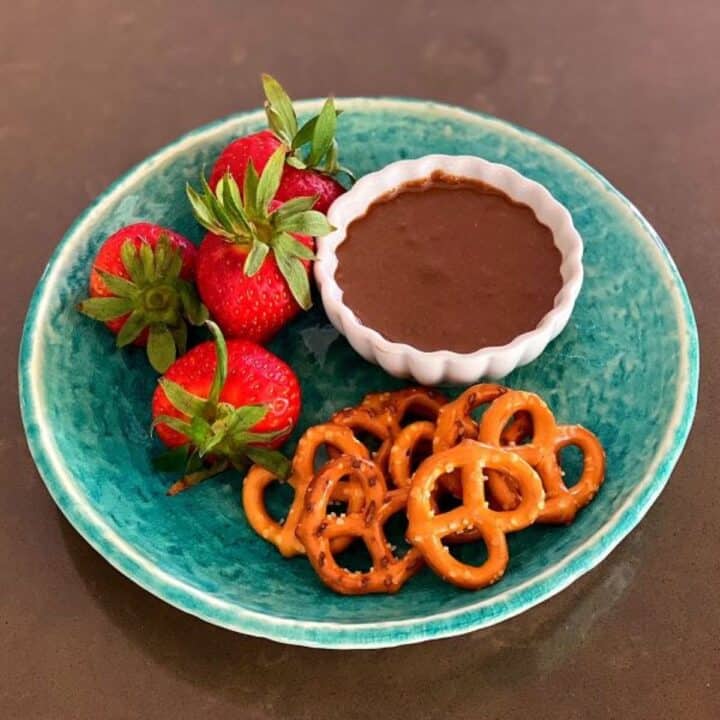 Chocolate pumpkin seed butter in small white ramekin with pretzels and strawberries on a blue-green plate.