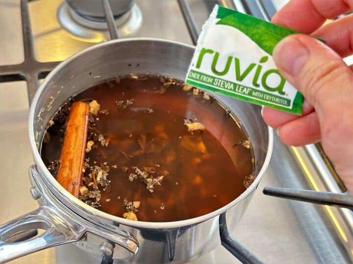 Pouring stevia from a green-white Truvia packet into ginger tea base in saucepan.