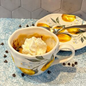 Porcelain tea cup decorated with lemons and filled with chocolate chip cake in a mug topped with whipped cream with saucer and spoon behind.