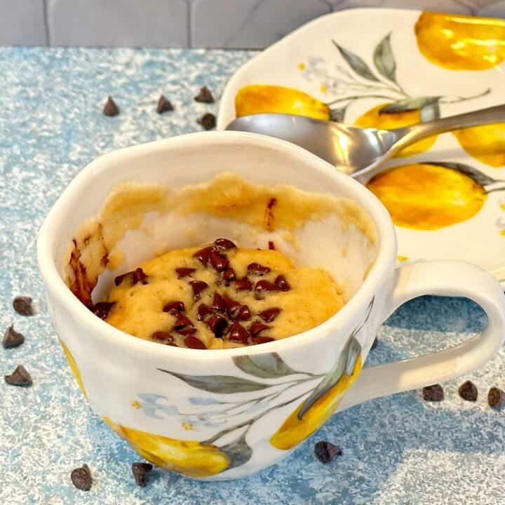 Porcelain tea cup with lemon motif filled with a chocolate chip cake. Lemon-motif porcelain saucer in background and mini chocolate chips scattered.