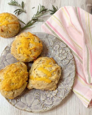 Three Rosemary Cheddar Biscuits on a floral stamped plate and one on a wooden countertop with fresh rosemary sprigs and pink striped napkin.