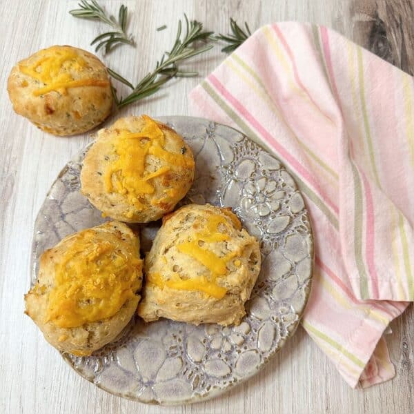 Three Rosemary Cheddar Biscuits on a floral stamped plate and one on a wooden countertop with fresh rosemary sprigs and pink striped napkin.