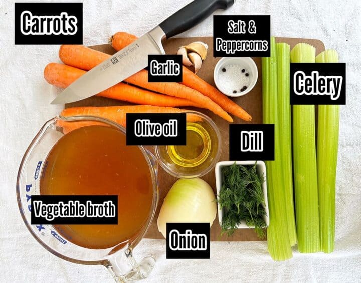 Broth, whole carrots and celery stalks, fresh dill, half an onion, garlic cloves, olive oil, salt and peppercorns with chef's knife on wooden cutting board.
