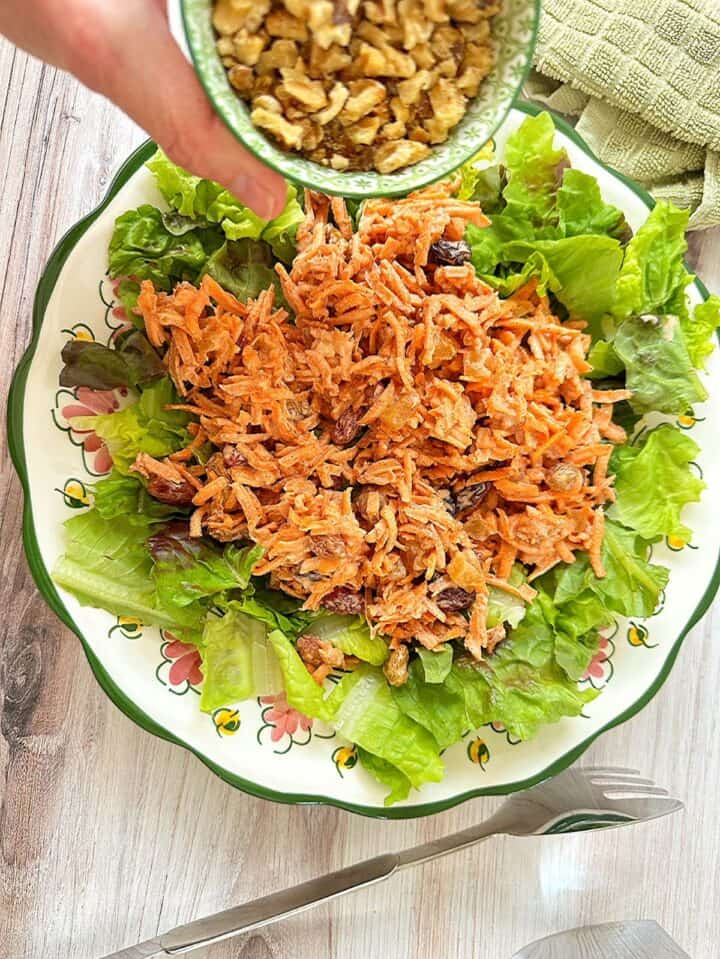 Shaking bits of toasted walnuts onto Carrot Pineapple Raisin Salad heaped on torn leafy lettuce on a large green-rimmed plate on a wood background.