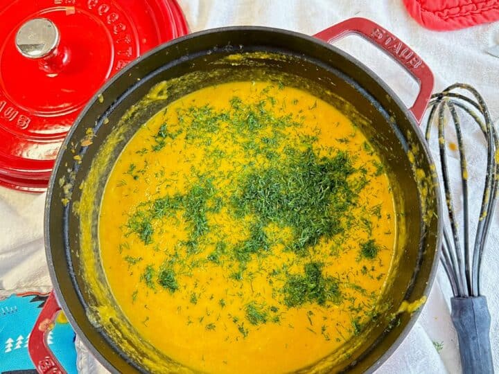 Creamy orange soup topped with lots of fresh dill in red Dutch oven with whisk and colorful potholders nearby on whitish background.