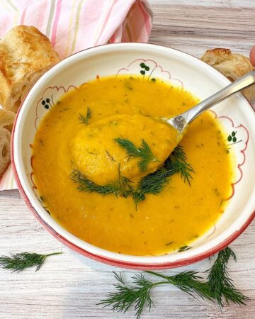 Creamy orange Carrot Celery Soup sprinkled with fresh, green dill with a spoon dipping in and a pieces of baguette and a pink cotton napkin aside it on a wooden background.