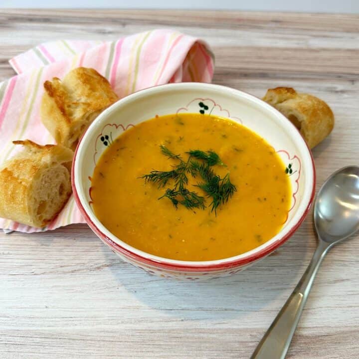 Creamy orange soup in pink-trimmed bowl topped with dill sprigs with a pink striped napkin, baguette slices, and spoon on a light wooden surface.