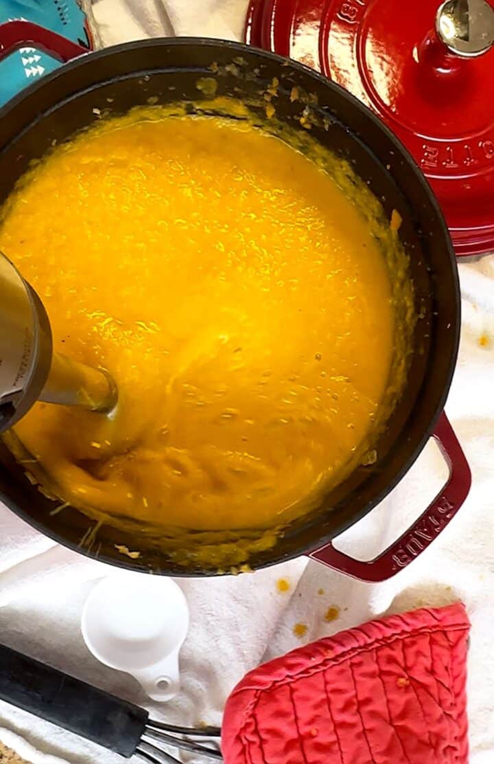 Immersion blender pureeing carrot and vegetables in red Dutch oven on a white background with red pot holder and pot lid.