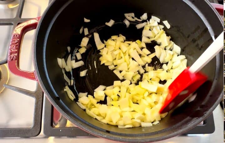 Onions cooking in a dark, heavy bottomed pot with red-headed spatula stirring.