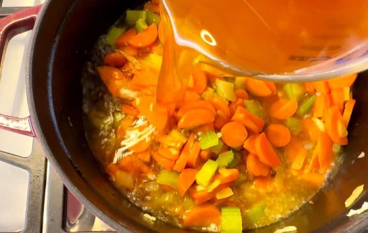 Pouring light brown broth into carrot-celery-onion mix in pot with dark interior or stovetop.