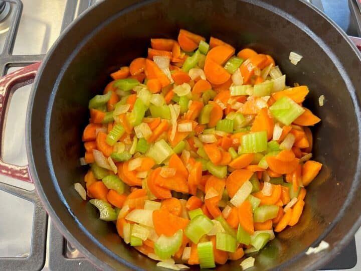 Carrot-celery-onion mix cooking in a red Dutch oven with dark interior on gas stovetop.