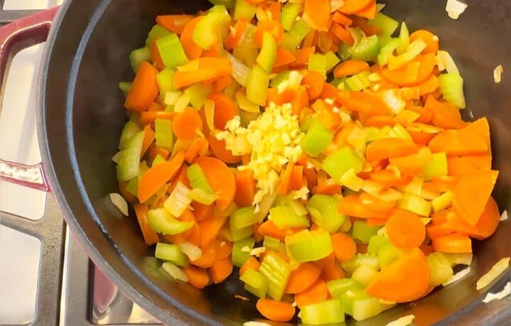 Softened carrot-celery-onion mix with minced garlic on top in a Dutch oven with dark interior on gas stovetop.