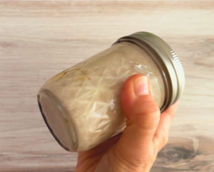 Hand holding a smallish quilted jam jar containing a creamy, off-white dressing with a wooden background.