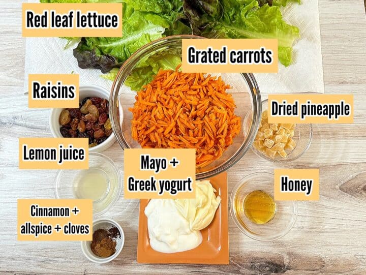 Grated carrots, dried pineapple, raisins, mayo, yogurt, and other ingredients in Carrot Pineapple Raisin Salad all with labels on wood grain background