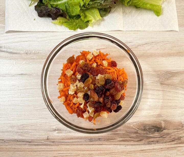 Clear bowl on wooden background holding grated carrots, dried pineapple bits, and raisins, with red leaf lettuce above.