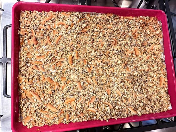 Granola with seeds and large toasted coconut shreds looking golden in a pink sheet pan on a stovetop surface.