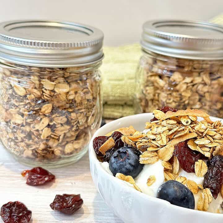 Fluted white bowl with blueberries and plain yogurt topped with granola, plus two small jars filled with granola and light green kitchen towel behind it with scattered dried cherries.