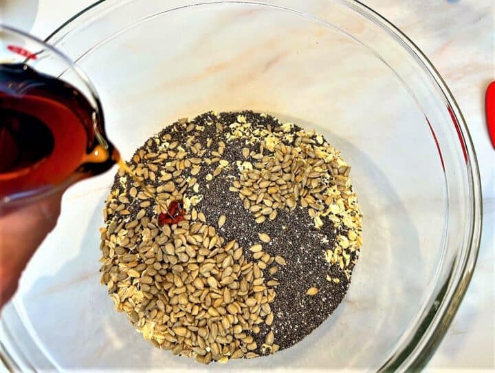 Maple syrup being poured into a clear bowl containing sunflower seeds, chia seeds, and oats,