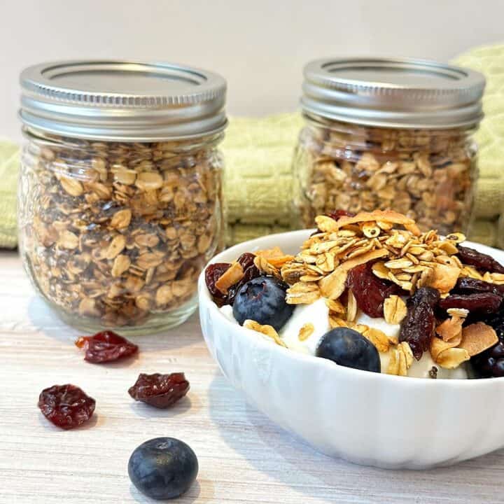 White bowl filled with blueberries, plain yogurt, and granola. Two small jars of granola in the back with green kitchen towel.