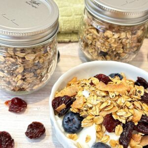 Fluted white bowl with blueberries and plain yogurt topped with granola and two jars of granola and a green kitchen towel behind it.