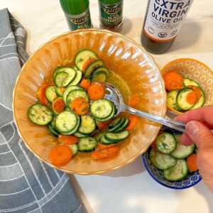 Slotted spoon dipping into a zucchini and carrot coin salad with kitchen towel. salt and pepper grinders, and olive oil in the background.