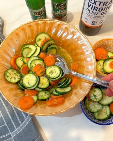 Slotted spoon dipping into a zucchini and carrot coin salad with kitchen towel. salt and pepper grinders, and olive oil in the background.