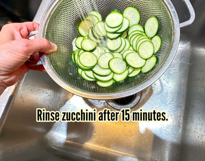Zucchini rounds in a steel colander being rinsed in a stainless steel sink.