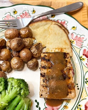 Tofu steak with sauce and grill marks, tiny roasted potatoes, and broccoli on pink and green pottery dinner plate.