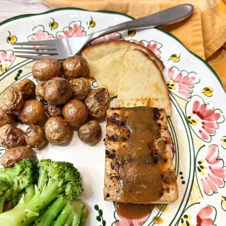 Tofu steak with sauce and grill marks, tiny roasted potatoes, and broccoli on pink and green pottery dinner plate.