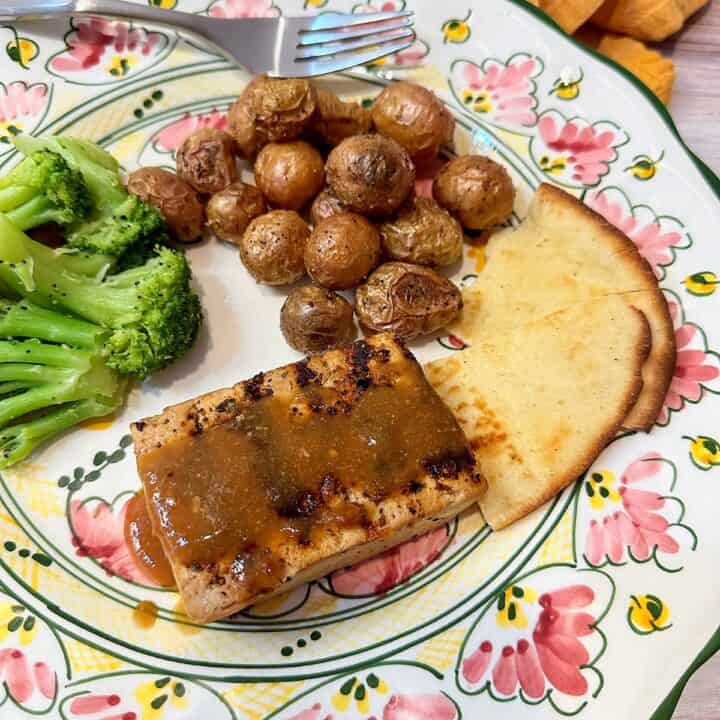 Tofu steak with grill marks and sauce and sides of baby potatoes and broccoli on a green-pink dinner plate