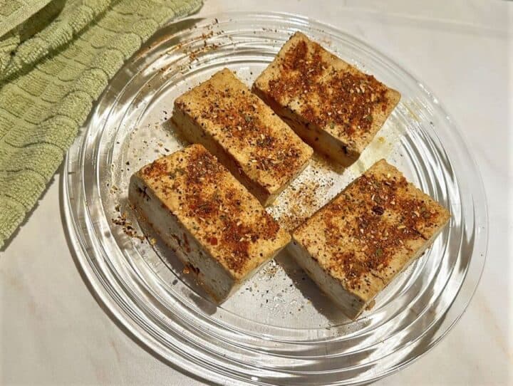 Four rectangular pieces of tofu coated with steak seasoning on a clear glass plate.