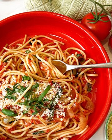 Shallow cherry red bowl with spaghetti coated in tomato sauce and topped with fresh basil and grated Parmesan.